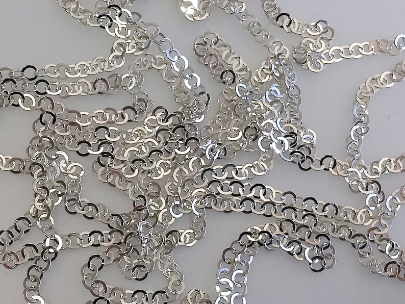 May include: A silver chain made of interlocking oval links. The chain is lying on a white surface.