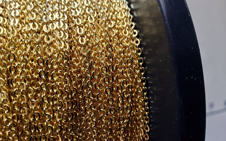 K&ouml;nnte beinhalten: Nahaufnahme einer Spule mit goldfarbener Kette. Die Kette besteht aus kleinen, miteinander verbundenen kreisf&ouml;rmigen Gliedern, die eine dichte, strukturierte Oberfl&auml;che bilden. Die Kette ist um eine schwarze Spule gewickelt.