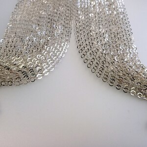 May include: A close-up of a silver chain necklace with a delicate, interwoven design. The chain is made of small, linked circles and is arranged in a flowing, layered pattern.