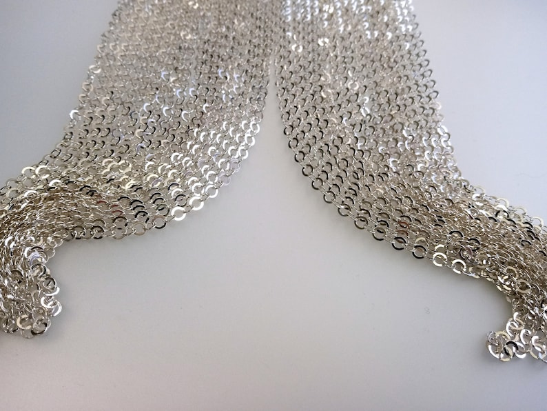 May include: A close-up of a silver chain necklace with a delicate, interwoven design. The chain is made of small, linked circles and is arranged in a flowing, layered pattern.