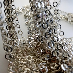 May include: A close-up of a silver chain, made of many small, interconnected rings. The chain is piled up in a heap, creating a textured and intricate pattern.