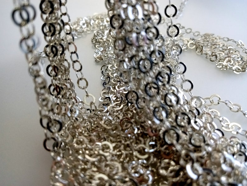May include: A close-up of a silver chain, made of many small, interconnected rings. The chain is piled up in a heap, creating a textured and intricate pattern.