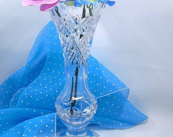 May include: A clear crystal vase with a diamond pattern holds pink and blue orchids. The vase sits on a white surface with a blue polka dot fabric in the background. The vase has a flared top and a round base.