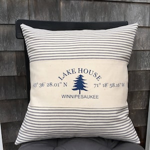 May include: A white and blue striped pillow with a white canvas insert. The canvas insert has a blue pine tree graphic and the text "LAKE HOUSE" in a blue arch above the tree. The text "43° 36' 28.01" N" is to the left of the tree and the text "71° 18' 58.16" W" is to the right of the tree. The text "WINNIPESAUKEE" is below the tree.