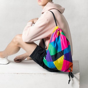 Peut inclure: Une jeune personne portant un sweat à capuche rose et un short noir est assise sur une surface blanche. Elle porte un sac à dos à cordon de serrage coloré avec une sangle noire.