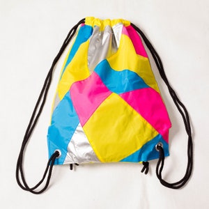 May include: A colourful drawstring bag made of patchwork fabric in shades of yellow, blue, pink, and silver. The bag has a cinched top and two long drawstrings.