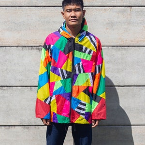 Puede incluir: Una persona lleva una chaqueta colorida con estampado geométrico, cremallera negra y capucha. La chaqueta tiene una variedad de colores, incluyendo rojo, amarillo, verde, azul y negro.