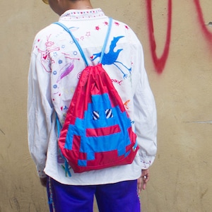 May include: A white button-down shirt with colorful drawings on it. The shirt has a blue drawstring backpack with a red and blue alien face on it.