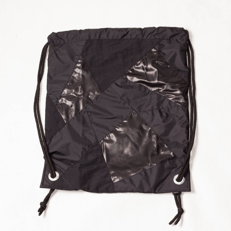 以下が含まれることがあります： 黒のパッチワーク生地で作られた黒い巾着袋。袋の下部に銀色のハトメが2つ付いています。