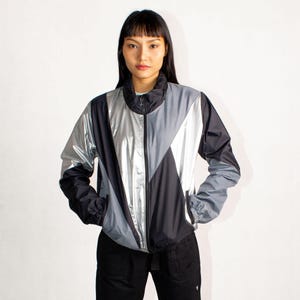 May include: A color-blocked jacket in black, silver, and gray. The jacket has a high collar, a full-length zipper, and two front pockets. The jacket is made of a shiny, lightweight material.