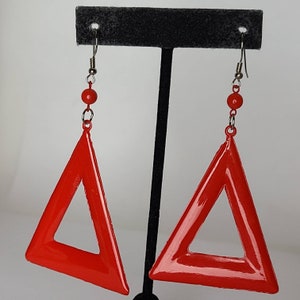 May include: A pair of red triangle earrings with a silver hook and a red bead. The earrings are hanging from a black stand.
