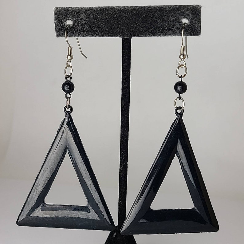 May include: A pair of black triangle earrings with silver hooks and black beads. The earrings are hanging from a black stand.