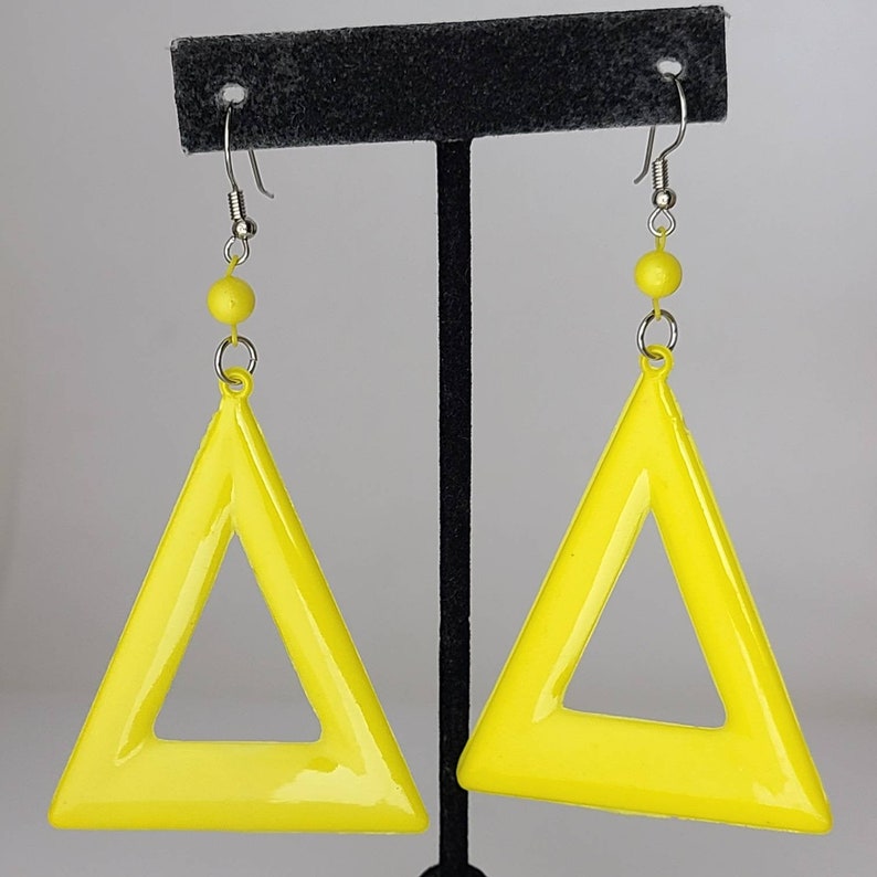 May include: A pair of yellow triangle earrings with a hollow center. The earrings are hanging from a black display stand.
