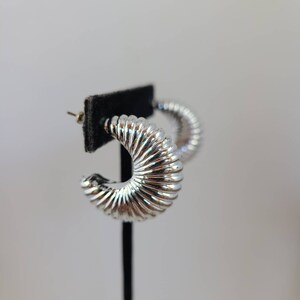 May include: Silver hoop earrings with a textured, ribbed design. The earrings are displayed on a black stand.