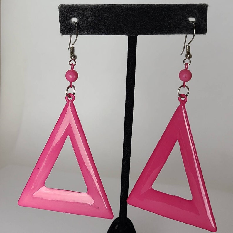 May include: A pair of pink triangle earrings with a hollow center. The earrings are hanging from silver hooks.