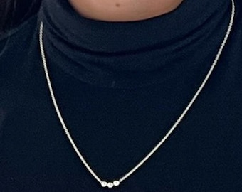 Peut inclure: Un collier en chaîne argent avec trois petites perles argentées qui pendent. Le collier est porté par une personne portant un pull à col roulé noir.