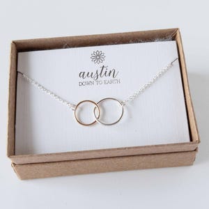 May include: A silver chain necklace with two interlocking circles, one gold and one silver. The necklace is in a brown box with a white card that says "austin down to earth".