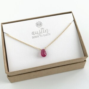 May include: A delicate gold chain necklace with a teardrop-shaped ruby pendant. The necklace is displayed in a white box with the text "austin down to earth".