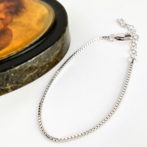 May include: A silver-toned box chain bracelet featuring a polished metallic finish, a secure lobster claw clasp, and an adjustable extension chain with small circular links, designed with a sleek and uniform square-link construction throughout.