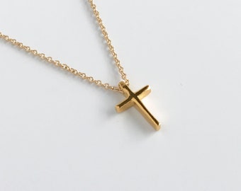 Pode incluir: Um pingente de cruz em metal dourado suspenso por uma delicada corrente de elos a condizer. A cruz apresenta uma superfície lisa e polida com arestas direitas e limpas, com um design simples e sem adornos. A corrente tem elos redondos.