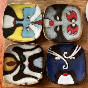 Peut inclure: Quatre petites assiettes carrées en céramique avec des motifs abstraits. Chaque assiette présente un motif unique aux couleurs jaune, bleu, noir et rouge. Les motifs sont symétriques et artistiques, avec une finition brillante. Les assiettes sont disposées sur une surface en bois.