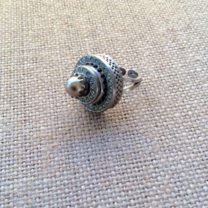 May include: A silver ring with a pearl-like center and a circular design of clear rhinestones.