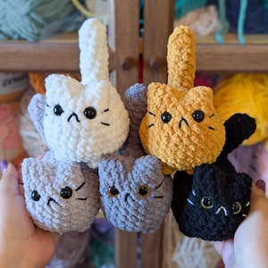 May include: A collection of handmade crochet cat toys in various colors, including white, gray, orange, and black. Each cat has black button eyes and embroidered facial features. The toys are held up against a backdrop of yarn.
