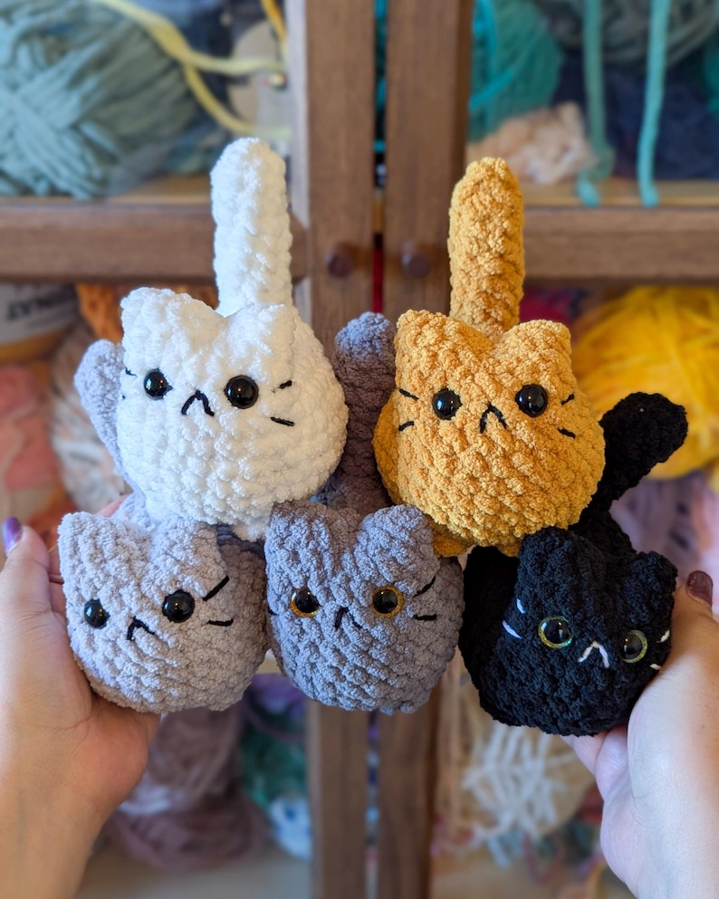 May include: A collection of handmade crochet cat toys in various colors, including white, gray, orange, and black. Each cat has black button eyes and embroidered facial features. The toys are held up against a backdrop of yarn.