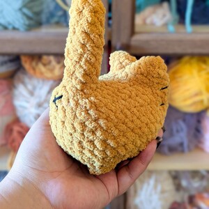 May include: A handmade, mustard-yellow crochet bunny toy. The plush bunny has a long, upright ear and a rounded body. Black thread details form the eyes and mouth. The soft, textured yarn gives the toy a cozy appearance.