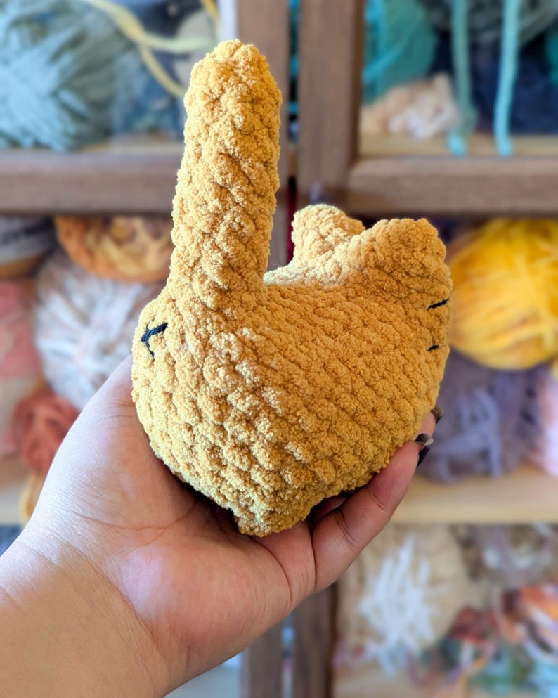 May include: A handmade, mustard-yellow crochet bunny toy. The plush bunny has a long, upright ear and a rounded body. Black thread details form the eyes and mouth. The soft, textured yarn gives the toy a cozy appearance.