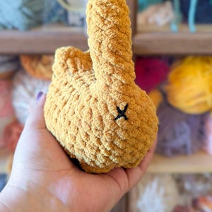 May include: A handmade, plush bunny in a warm yellow hue. The bunny has a textured, fuzzy appearance and a small black 'X' stitched on its rear. The image shows the bunny being held, with colorful yarn in the background.