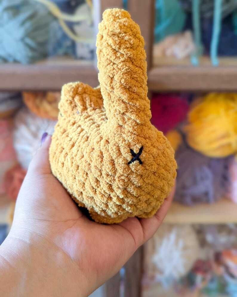 May include: A handmade, plush bunny in a warm yellow hue. The bunny has a textured, fuzzy appearance and a small black 'X' stitched on its rear. The image shows the bunny being held, with colorful yarn in the background.