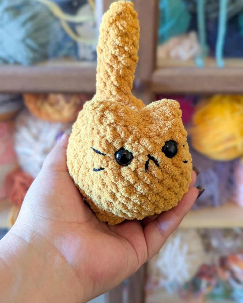 May include: A handmade, golden-yellow crocheted cat plushie with black eyes and whiskers. The cat has a small, upright tail and is held in a hand. The background shows yarn in various colors.