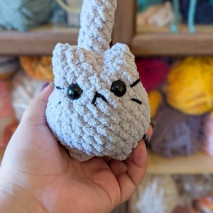 May include: A handmade, light gray crocheted cat toy with black eyes and whiskers. The cat has a rounded body and a long, upright tail. The toy is held in a person's hand, with yarn in the background.