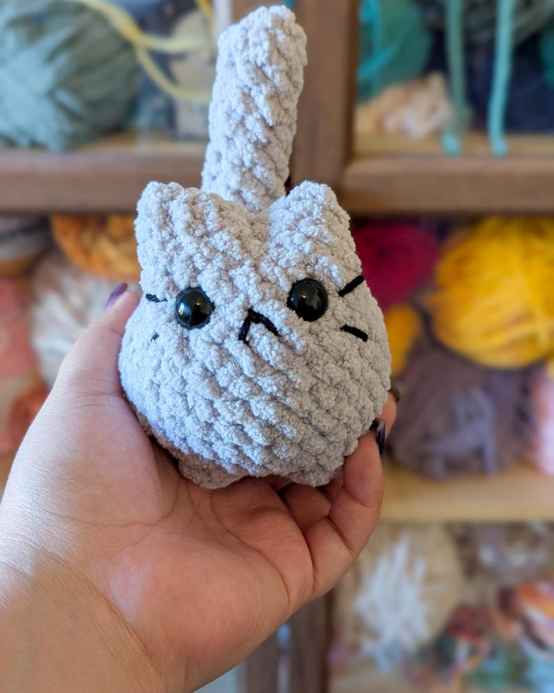 May include: A handmade, light gray crocheted cat toy with black eyes and whiskers. The cat has a rounded body and a long, upright tail. The toy is held in a person's hand, with yarn in the background.