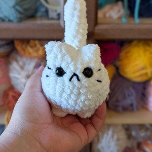 May include: A handmade white crochet cat with black eyes and whiskers. The cat has a fluffy tail and a grumpy expression. The cat is made of soft yarn and is a cute, whimsical item.
