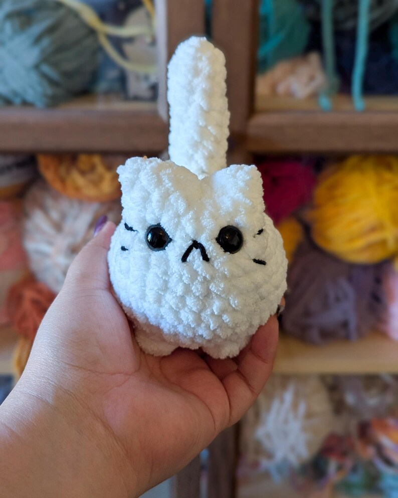May include: A handmade white crochet cat with black eyes and whiskers. The cat has a fluffy tail and a grumpy expression. The cat is made of soft yarn and is a cute, whimsical item.
