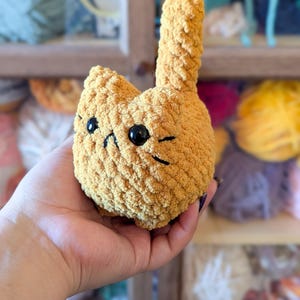 May include: A handmade, golden-yellow crochet cat plushie with black eyes and whiskers. The cat has a rounded body and a tall, upright tail. The plushie is held in a hand, showcasing its soft, textured yarn construction. A charming, whimsical item.
