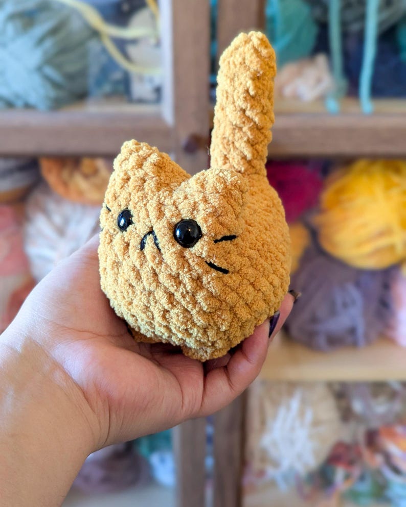 May include: A handmade, golden-yellow crochet cat plushie with black eyes and whiskers. The cat has a rounded body and a tall, upright tail. The plushie is held in a hand, showcasing its soft, textured yarn construction. A charming, whimsical item.