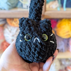 May include: A handmade black crochet cat with large, glittery green eyes and white whiskers. The cat has a small, upright tail. The plush toy is held in a hand, with a background of colorful yarn.