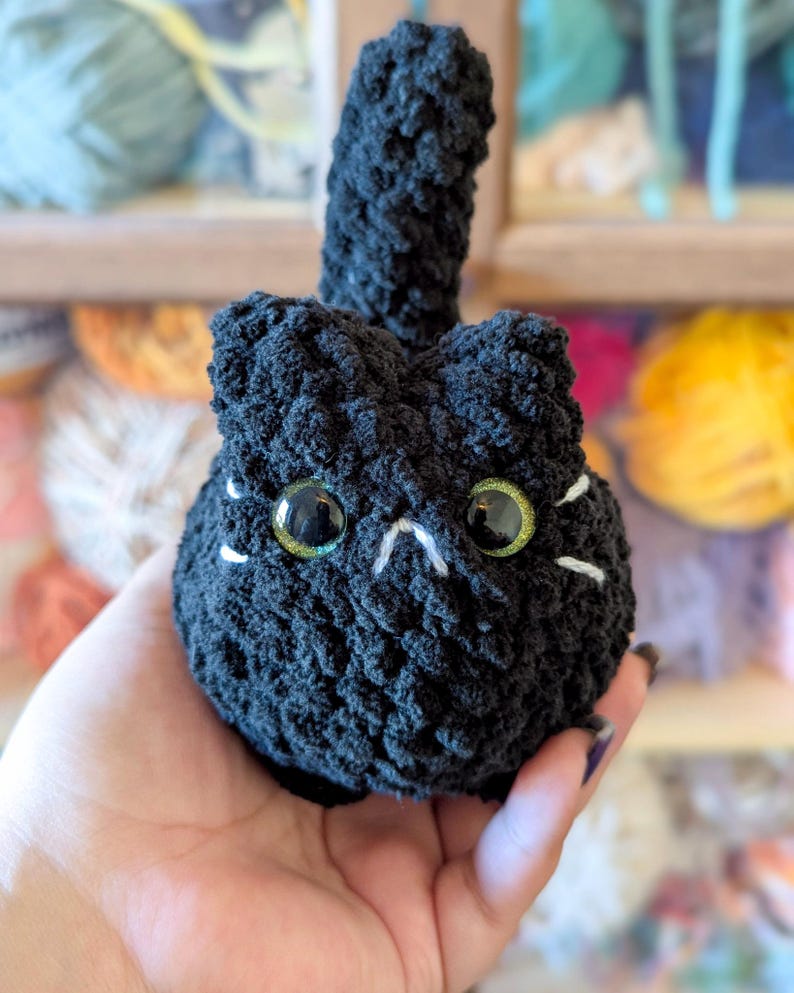 May include: A handmade black crochet cat with large, glittery green eyes and white whiskers. The cat has a small, upright tail. The plush toy is held in a hand, with a background of colorful yarn.