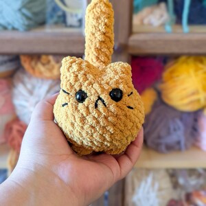 May include: A handmade, golden-yellow crocheted cat toy with black eyes and whiskers. The cat has a small, upright tail. The toy is held in a hand, with a blurred background of yarn skeins.