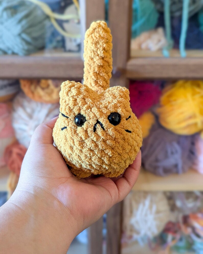 May include: A handmade, golden-yellow crocheted cat toy with black eyes and whiskers. The cat has a small, upright tail. The toy is held in a hand, with a blurred background of yarn skeins.