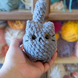 May include: A handmade gray crochet cat with large golden eyes and black whiskers. The plush cat has a textured surface and a raised tail. The cat is a soft, cuddly toy.
