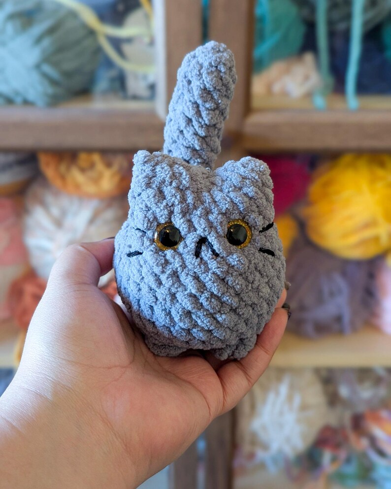 May include: A handmade gray crochet cat with large golden eyes and black whiskers. The plush cat has a textured surface and a raised tail. The cat is a soft, cuddly toy.