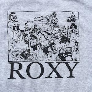Könnte beinhalten: Graues T-Shirt mit einem schwarz-weißen Grafikdruck einer Gruppe von Menschen in einer Szene mit dem Wort "ROXY" unterhalb der Grafik.