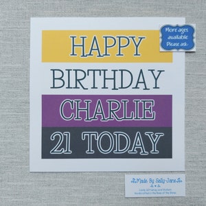 May include: A white card with black text that reads "Happy Birthday Charlie 21 Today". The card has three coloured stripes: yellow, purple, and grey. A small blue sticker with white text reads "More ages available Please ask".