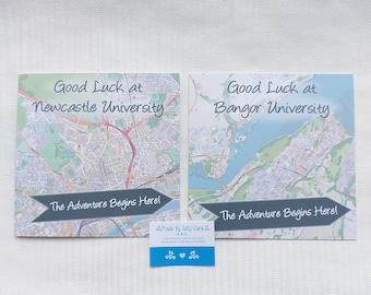 May include: Two cards with maps of Newcastle University and Bangor University. Each card has the text "Good Luck at [University Name]" and "The Adventure Begins Here!" printed on them. The cards are on a white surface.