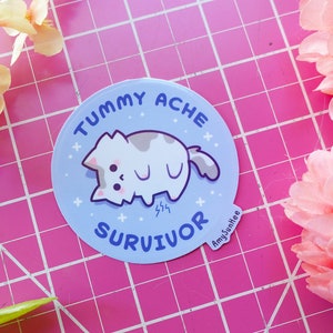 May include: A blue and white sticker with a cartoon cat curled up on its side. The sticker says "Tummy Ache Survivor" with a small star design around the edge. The sticker is on a pink and white grid background.