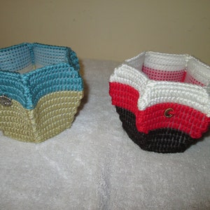 May include: Two handmade, hexagonal-shaped baskets. One is teal and beige with a silver embellishment, the other is red, white, and black with a gold embellishment. The baskets are made of a woven material.
